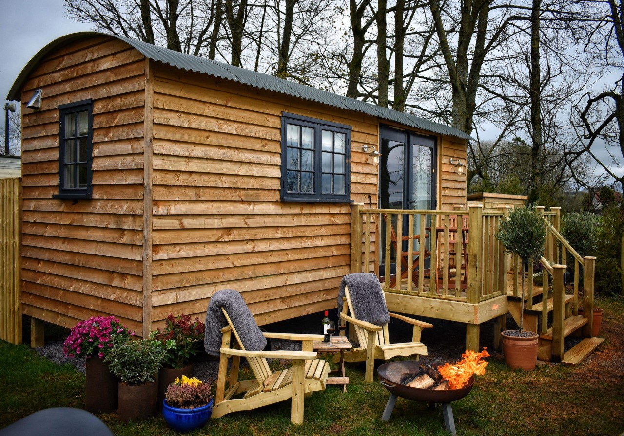 A luxury shepherds hut with a curved roof sits in a wooded area. Two wooden chairs with grey blankets are positioned around a fire pit on a deck, surrounded by potted plants and flowers. A bottle of wine and two glasses are on a small table between the chairs. Perfect accommodation for two near Alton Towers