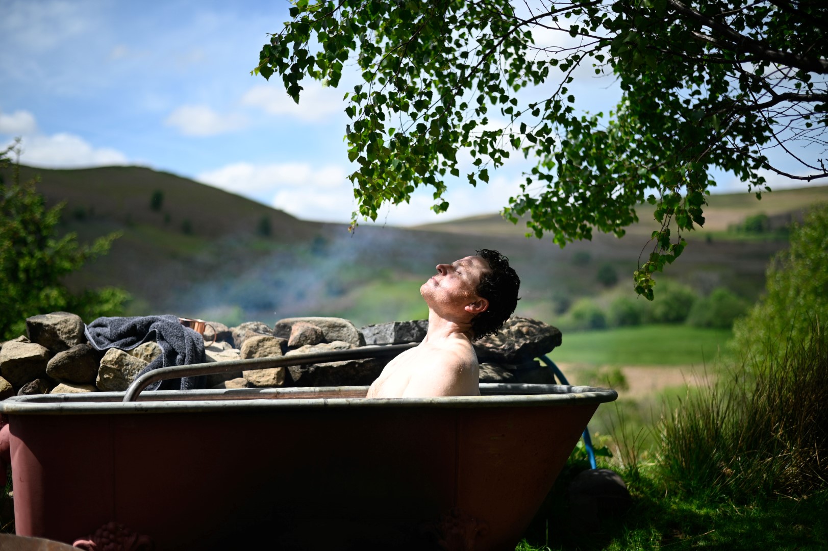 A person relaxes in an outdoor bathtub set among rocks, enjoying the sunshine. Overhead, a tree branch with green leaves adds a natural canopy. The background features rolling hills and a partly cloudy sky—bask in serene UK stays with outdoor baths. A towel is draped over the edge of the tub. Chillderness Red Kite Glamping